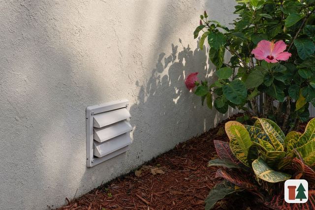 Louvered dryer vent hood on a stucco wall beside a hibiscus